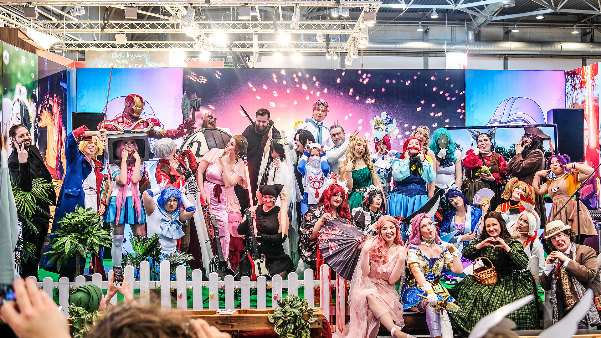 Cosplayer in farbenfrohen Kostümen posieren lachend auf der Bühne des Elbenwald-Stands bei der Leipziger Buchmesse 2025, umgeben von Deko, Pflanzen und einem leuchtenden Hintergrund mit Feuerwerkseffekt.
